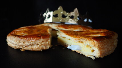 French puff pastry and almond cream Kings Cake, or Galette des Rois, crown on top, a slice cut showing the charm inside, on black background, horizontal picture