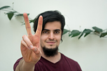 Young caucasian black haired man with beard and red shirt looking at camera with his hand on the front
making a peace hand sign or symbol. White background and climbing plant with leaves behind