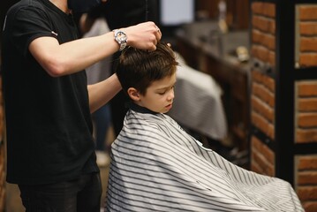 A little cute boy sits in a hairdresser's at the stylist's, a schoolchild is getting hair cut in a beauty salon, a child at a barbershop's, a short men's haircut.