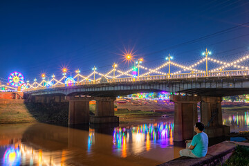 Fototapeta na wymiar The young man looking at Beautiful light on the Nan River at night on the bridge (Naresuan Bridge) in Realm for Naresuan the Great Festival and Red Cross Annual event in Phitsanulok,Thailand.