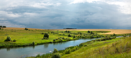 Summer cloudy landscape with dark stormy clouds over the calm river and green meadows.