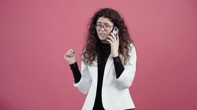 Agitated young woman with curly hair, talking on the phone with contempt gesturing with her hand. Young hipster in white jacket and black shirt, with glasses posing isolated on pink background in the