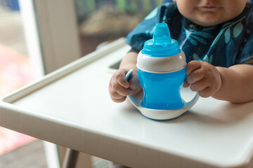 Breast fed baby learning to use a sippy cup to drink water; hands grasping handles of a blue plastic cup