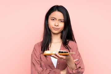 Teenager Asian girl eating sushi isolated on pink background keeping arms crossed