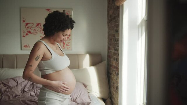 Pretty young Pregnant Woman Stands In Front Of Window And Touchs Belly. Happy woman smiling and looking at stomach. Medium shot.