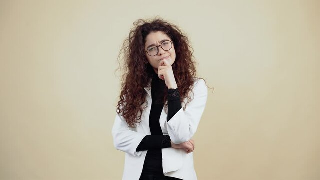 The young woman inspired by curly hair, keeps her finger on her lips and thinks of a solution. Young hipster in gray jacket and white shirt, with glasses posing isolated on beige background in the