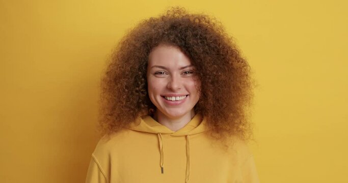 Positive good looking curly haired millennial girl shows goodbye gesture waves palm at camera and smiles gladfully dressed in casual sweatshirt isolated over yellow background. Nice to meet you
