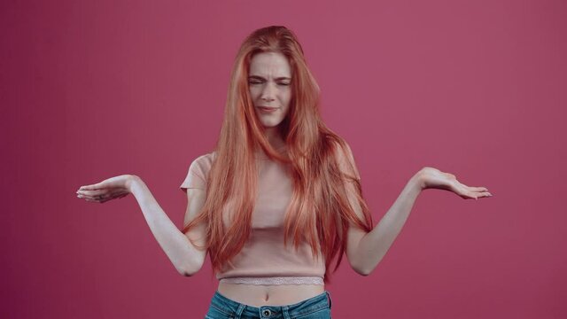 The puzzled redhead, bent over, does not understand, and asks once more, dissatisfied at the end. 20-year-old freckled hipster teenager in a pink casual T-shirt, isolated on a pink background.