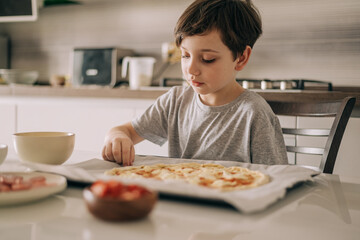 Little kid boy making pizza sitting at the table on the kitchen. Children helping in cooking lifestyle image