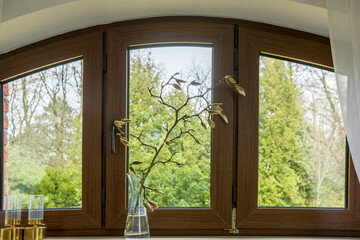 Large oval window with green trees in the background. A small magnolia branch with buttons is in a vase on the windowsill. Decoration in spring 