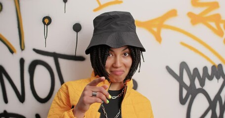 Cool looking hipster girl looks with wondered expression reacts on something then smiles cheerfully hears something funny dressed in fashionable clothes poses against colorful graffiti wall.