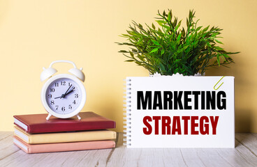 MARKETING STRATEGY is written in a notebook next to a green plant and a white alarm clock, which stands on colorful diaries.