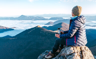 Young hiker female sitting on the mountain summit cliff and enjoying mountains valley covered with...