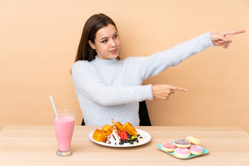 Teenager girl eating waffles isolated on beige background pointing finger to the side and presenting a product