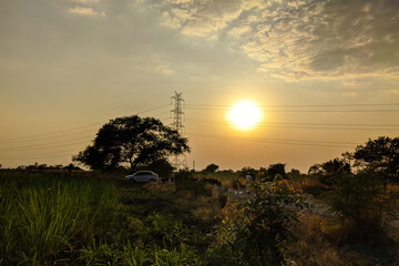 Dark shot of a car parked next to a tree and a cell tower during a cloudy sunset in the countryside