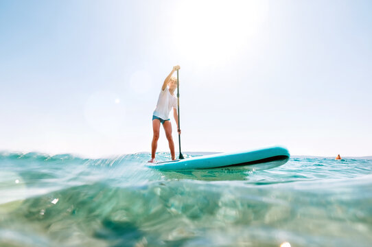 Under the water view angle to the smiling blonde teenager boy rowing stand up paddle board. Active family summer vacation time near the sea concept image.
