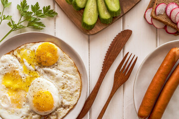 Fried eggs, fresh vegetables, smoked sausages for breakfast on wooden background.