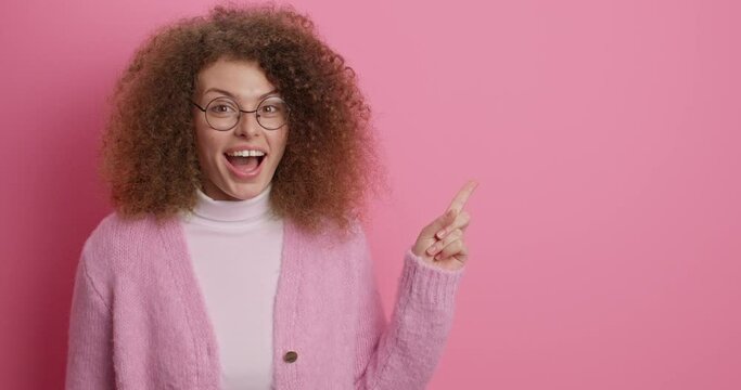 Read this information. Positive mysterious curly woman makes secret gesture tells confidential information shows best promotional deal shopping link or website logo dressed casually isolated on pink