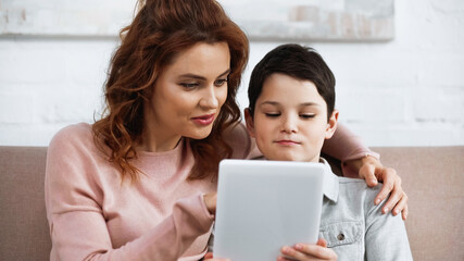 Smiling woman hugging son and using digital tablet during online education.
