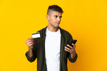 Young brazilian man isolated on yellow background buying with the mobile with a credit card while thinking