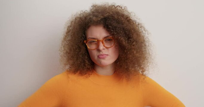 Pissed off outraged curly haired young woman shakes fist and threatens promises to revenge looks angrily at camera stands frustrated wears spectacles orange jumper isolated over white background
