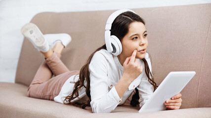 Pensive preteen girl in headphones holding digital tablet on couch at home.