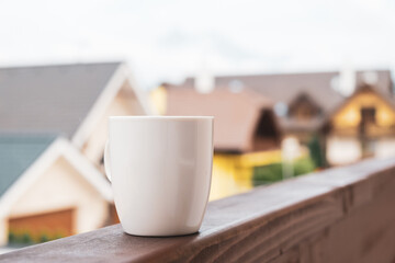 The steam rises from a cup of hot coffee or tea on the balcony with mountains and roofs of houses on the background. 