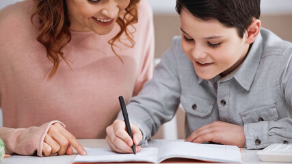 Smiling kid writing on notebook near mother pointing with finger.