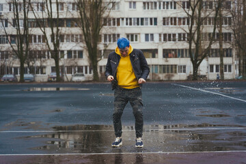 Young man jumps in puddle on sports stadium on background of high-rise building. Happy guy with blue hair having fun on city street in springtime.