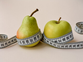 A pear and an apple are wrapped with a tape of a centimeter, a beige background. Close-up. Healthy food and lifestyle theme.