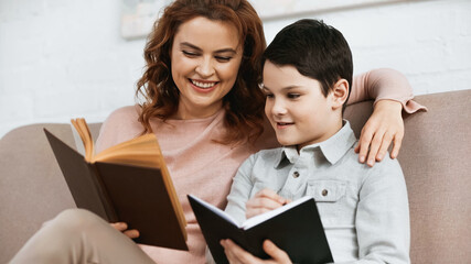 Cheerful woman reading book while helping son with homework.