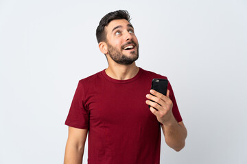 Young handsome man with beard using mobile phone isolated on white background looking up while...
