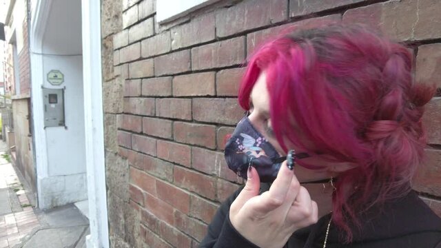 Young woman with pink red hair using or putting on a mask for medical protection against virus or bacteria. Street brick wall on the background