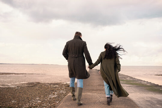 Couple in winter coats holding hands on ocean beach jetty