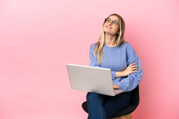 Young woman sitting on a chair with laptop over isolated pink background looking up while smiling