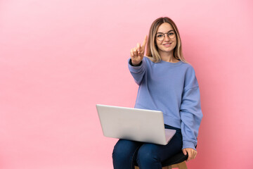 Fototapeta premium Young woman sitting on a chair with laptop over isolated pink background showing and lifting a finger