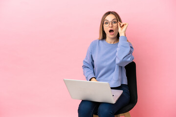 Young woman sitting on a chair with laptop over isolated pink background with glasses and surprised