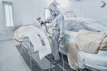 Car painting and automobile repair service. Auto mechanic in white overalls paints car with airbrush pulverizer in paint chamber.