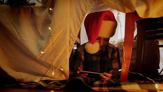 Little boy in Santa hat in DIY kid's tent laughing celebrating Christmas using smartphone, having joyful online conversation. Christmas fun at home.