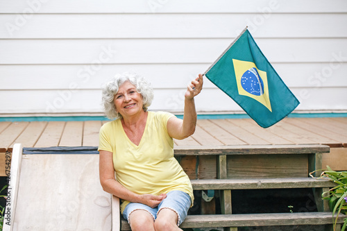 Proud citizen celebrating Independence Day of Brazil. Happy old woman with Brazilian flag outdoor. Smiling Brazilian elderly lady sitting on home backyard waving a Brazilian flag.