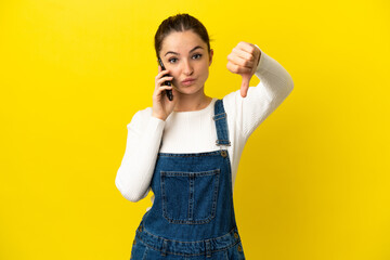 Young woman using mobile phone over isolated yellow background showing thumb down with negative...