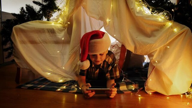 Happy little boy in Santa hat lying in DIY child tent celebrating Christmas using smartphone, having video call. Christmas night, home entertainment, long distance communication concept.