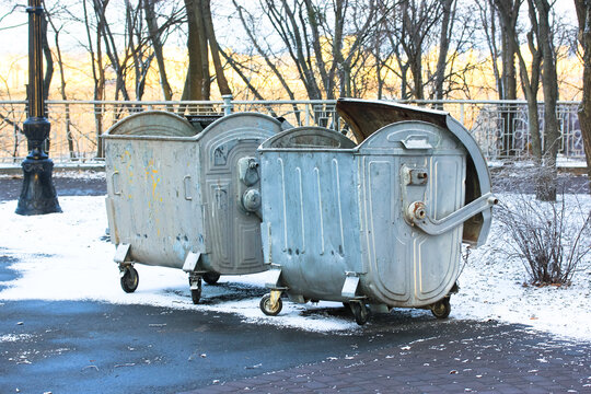 Large metal garbage cans, recycling bins, trash containers on wheels outside in the winter snowy park at daytime.
