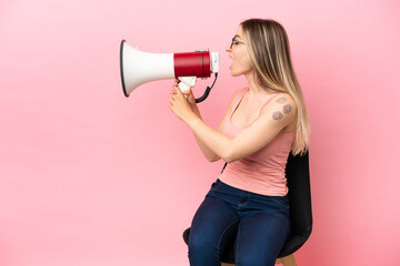 Young woman sitting on a chair over isolated pink background shouting through a megaphone