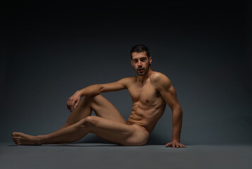 Obraz premium Gorgeous naked male model sitting isolated on the floor in a studio while looking in the camera
