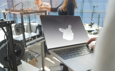 The electronics development engineer looks at the 3D rendering on the laptop next to the 3D printer while the other engineers prepare the 3D printer in the background