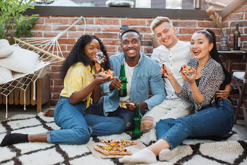 Cheerful mixed race friends in stylish clothes enjoying delicious pizza and alcoholic drinks while resting on roof top. Young men and women smiling and looking at camera.