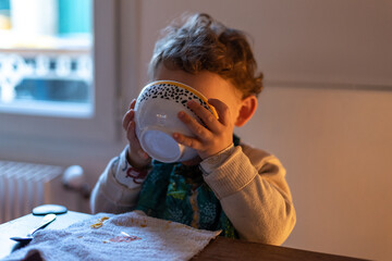 Cute caucasian child with blond curly hair drinking a soup while holding his bowl with both hands.