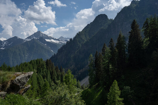 Wonderful view of Alpe Cheggio. Snow-capped mountains in the background, trees, blue sky, clouds. On the left, a group of rocks protrudes.