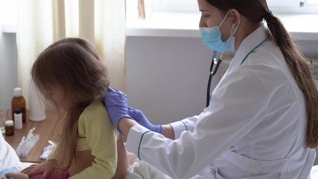 authentic cute caucasian cute young woman nurse or doctor pediatrician examine with statoscope minor preschool baby girl kid on white bed in hospital. medicine and health, pediatrics, covid-19 concept